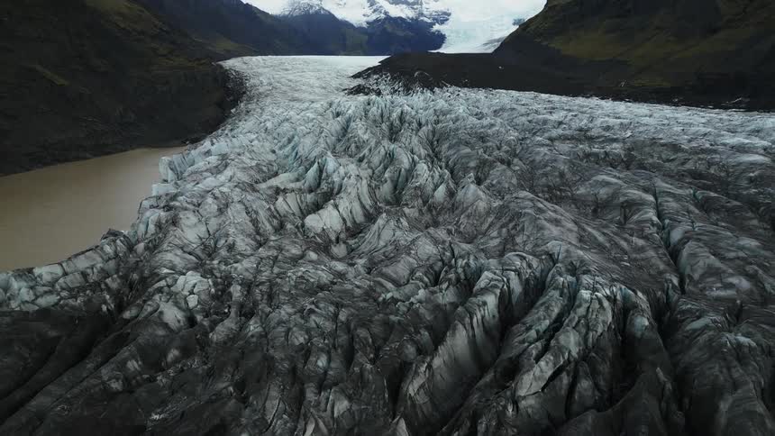 A textured glacier tongue descends between dark rocky mountains leading to a still lake with a muddy bank all under an overcast sky