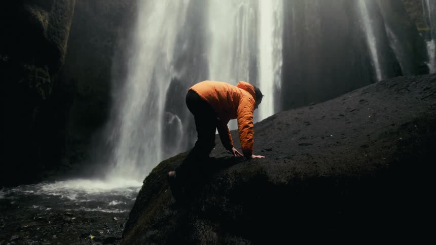 A person in an orange jacket is climbing a large rock near the base of a powerful waterfall with mist and water cascading down the cliffside in the background
