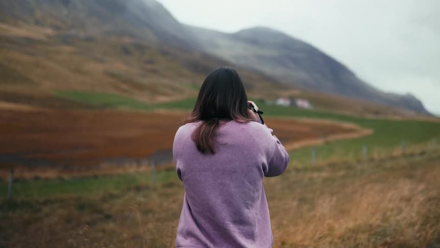 Woman from behind photographing a natural scene of a field with imposing mountains in the background