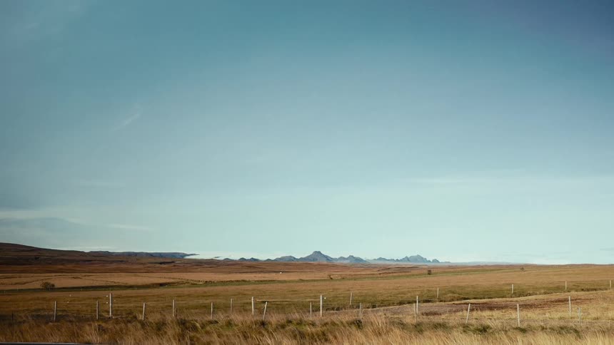 A landscape of a field with beige grass and mountains in the background and a white car passes in front