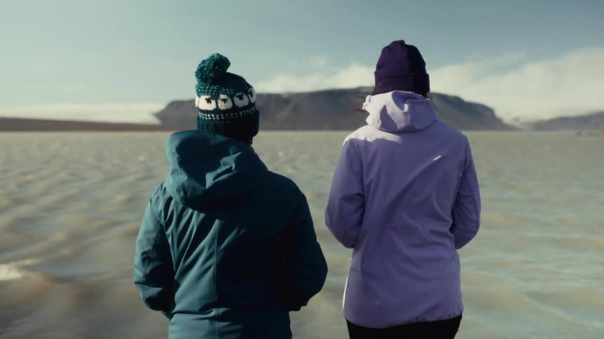 Two people wearing winter jackets and hats are standing side by side facing a large calm lake with a mountainous landscape in the background