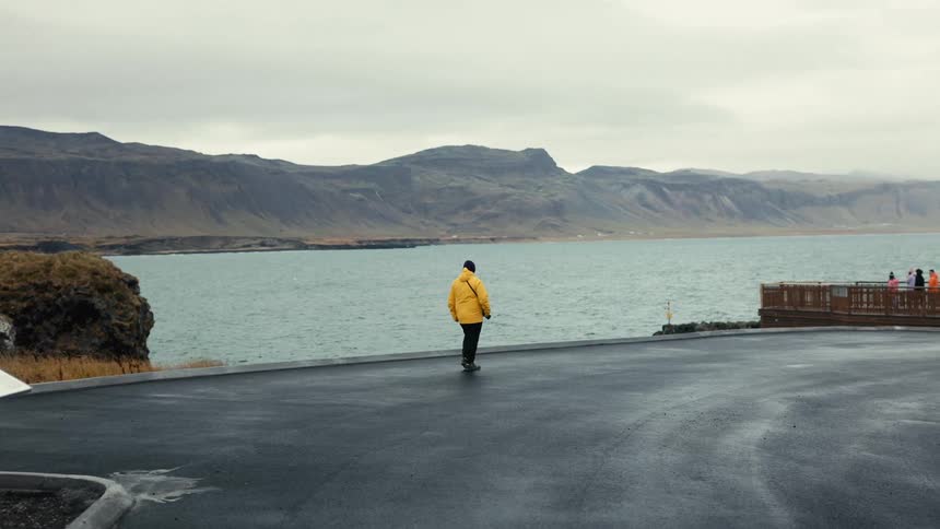 A person with their back turned wearing a yellow coat and walking on a concrete surface in front of a large body of water with mountains in the background