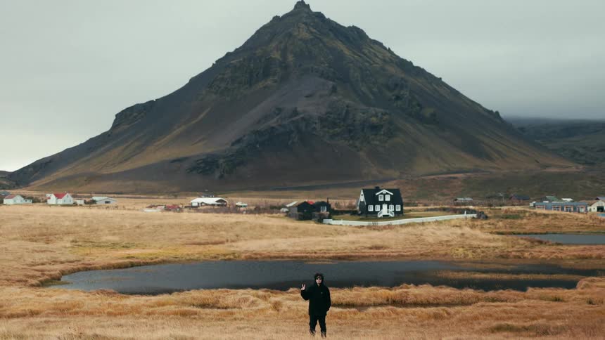 A person dressed in black stands in a vast golden field with a small pond in the foreground a village in the middle ground and a large rugged mountain dominating the background under a cloudy sky