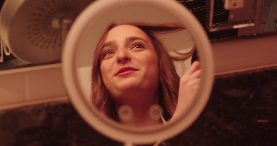 A teenage girl is using a curling iron to style hair as seen through a round illuminated vanity mirror in a bathroom setting