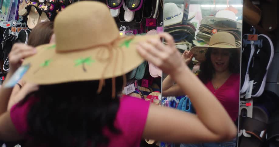 Woman next to her friend having fun trying on a hat in front of the mirror in a store