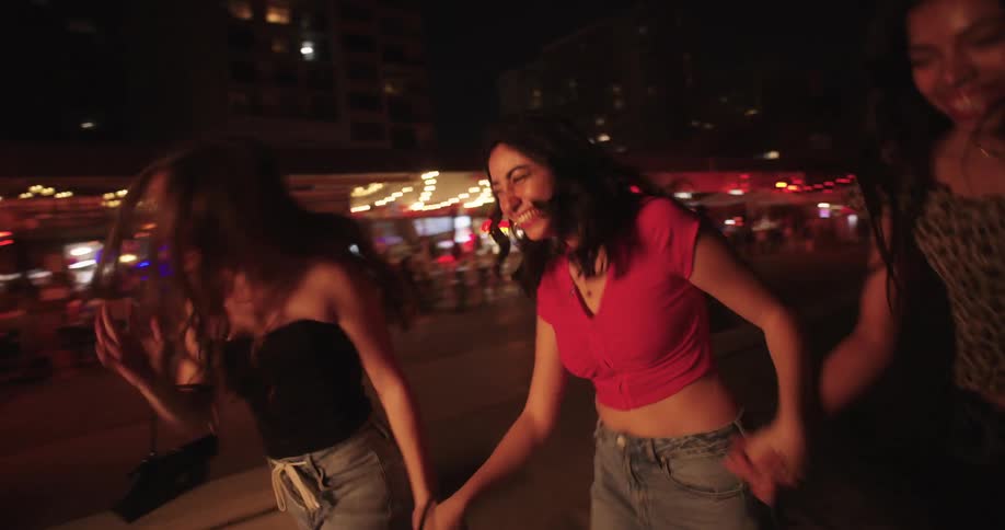 Three happy female friends running outdoors at night around bars