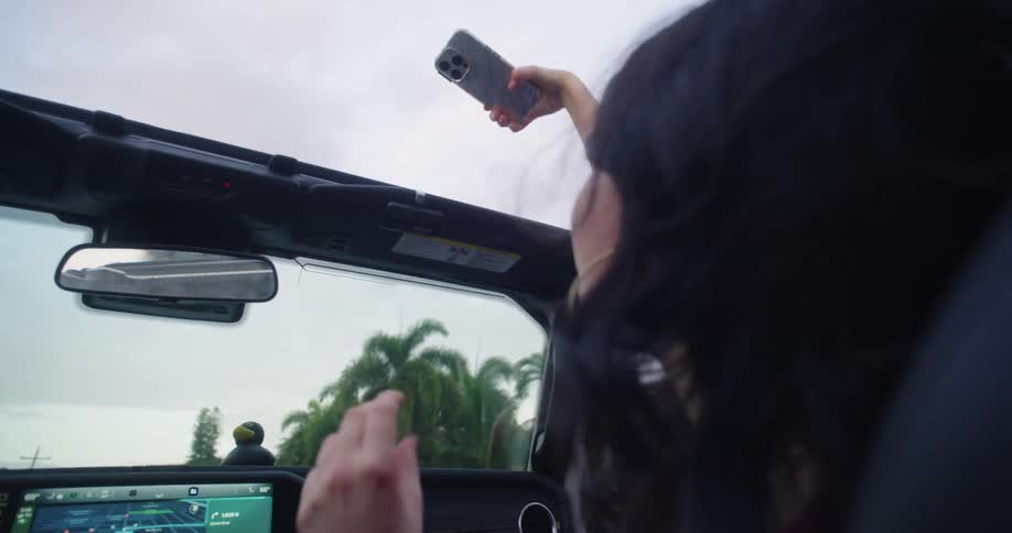 A teenage girl is holding a smartphone in a moving convertible car capturing a video of herself and a friend dancing