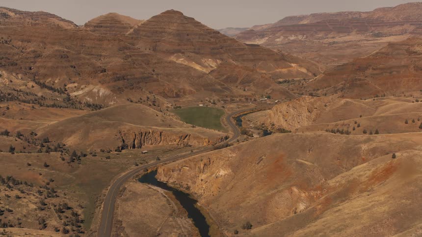 An aerial view shows a rural road running parallel to a river through a valley surrounded by hills and mountains