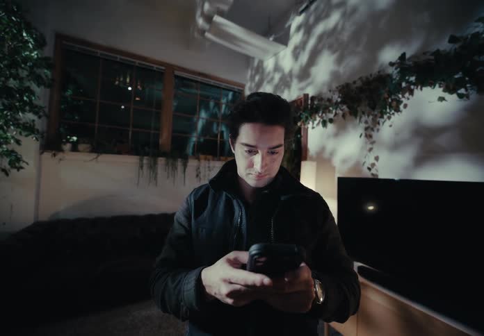 A young man in a dimly lit modern living space focused on his smartphone with soft lighting casting shadows on the walls surrounded by greenery and a large window in the background