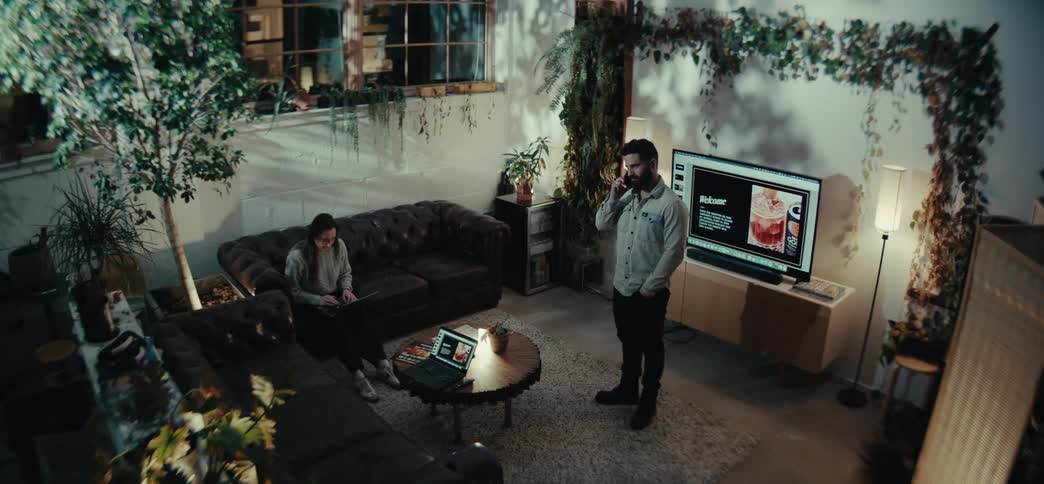 A modern living room with lush indoor plants a person sitting on a dark leather couch using a laptop another person standing and talking on a phone and a television displaying a welcome screen