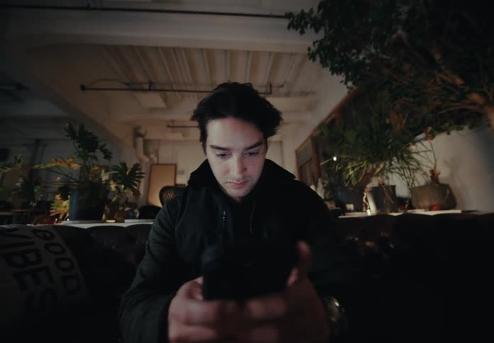A man in a black jacket sits on a dark leather couch and looks at his phone texting