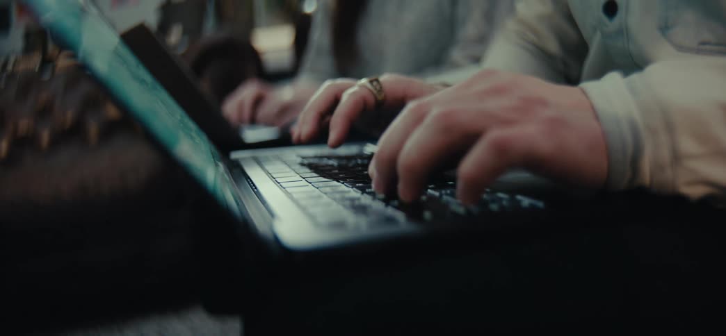A closeup shows hands typing on a laptop keyboard with another person blurred in the background also using a laptop