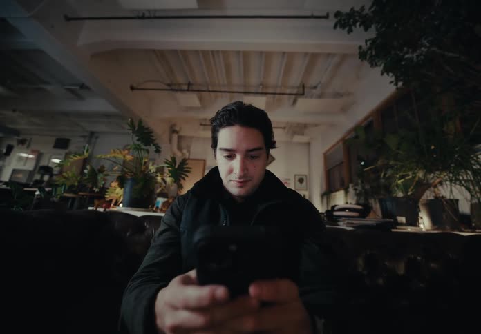 A person is sitting on a dark brown leather couch in a dimly lit room filled with various plants and decorations texting on a smartphone with both hands