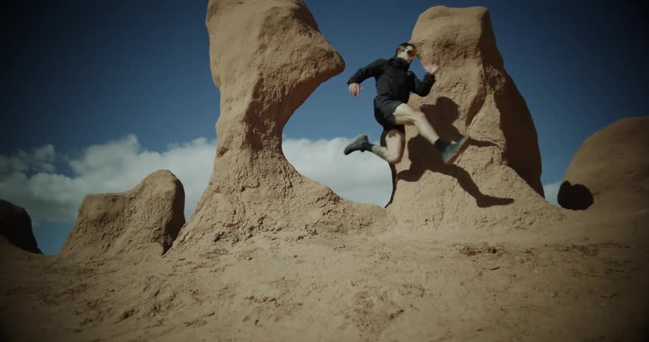 A man in a black jacket dark shorts and sunglasses leaps off a rock formation in a desert landscape