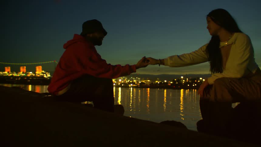 Silhouetted against a twilight cityscape and water reflections a man and a woman sit facing each other and holding hands