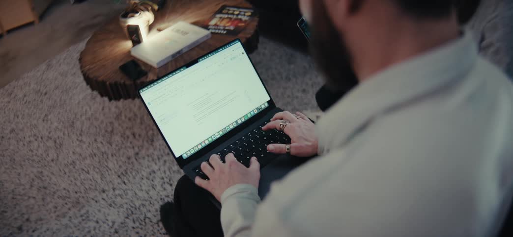 A person types on a laptop while sitting on a carpeted floor next to a wooden table with a book and magazines on it