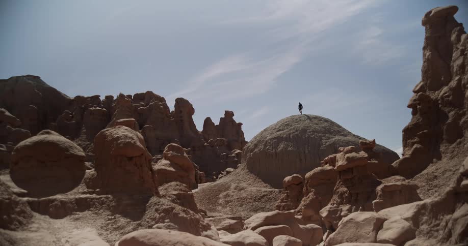A person stands atop a large rounded rock formation in a desert landscape filled with unique eroded sandstone structures under a partly cloudy sky