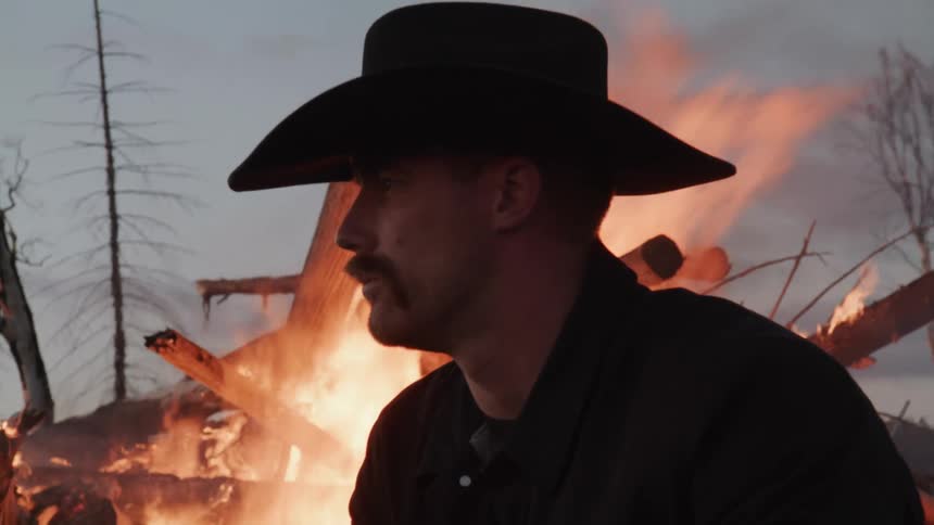 Man in hat outdoors with tree trunks on fire in background