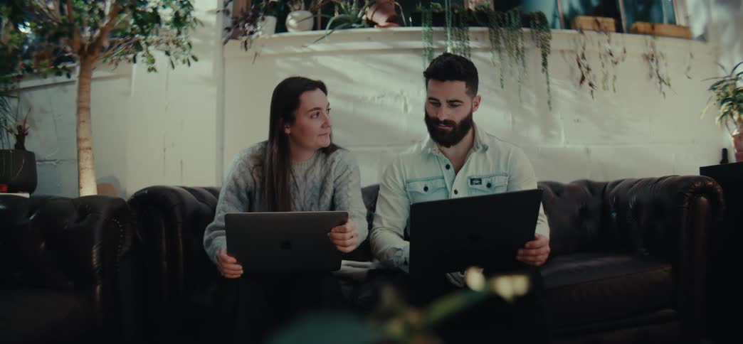 Two people sit on a dark leather couch working on laptops on a modern living room