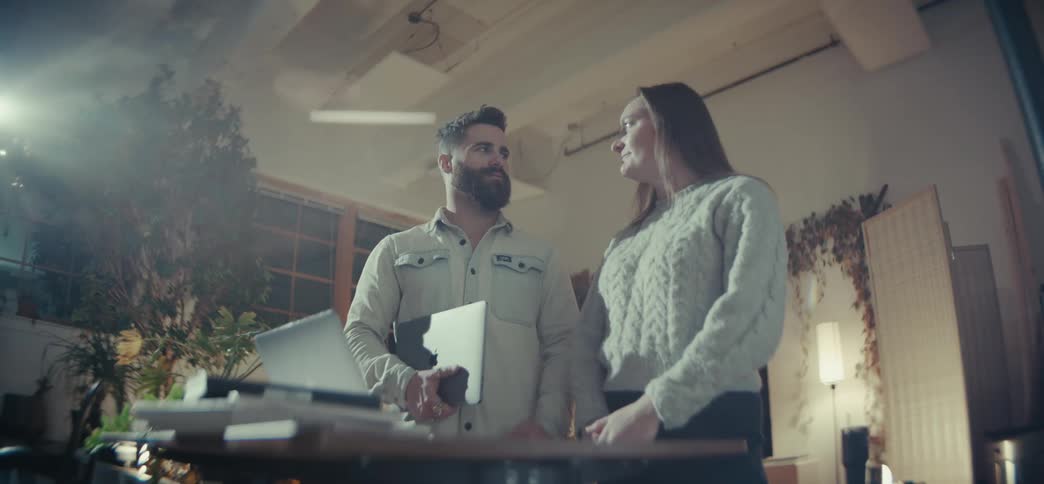 Two people are standing in an officelike environment one holding a laptop with various plants and a lamp in the background