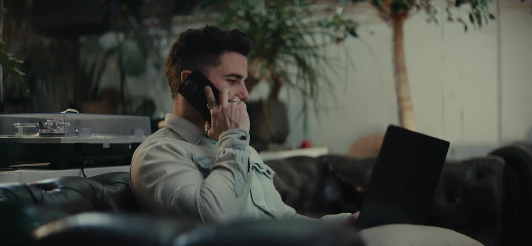 A man sits indoors on a sofa while answering his smartphone and typing on his laptop