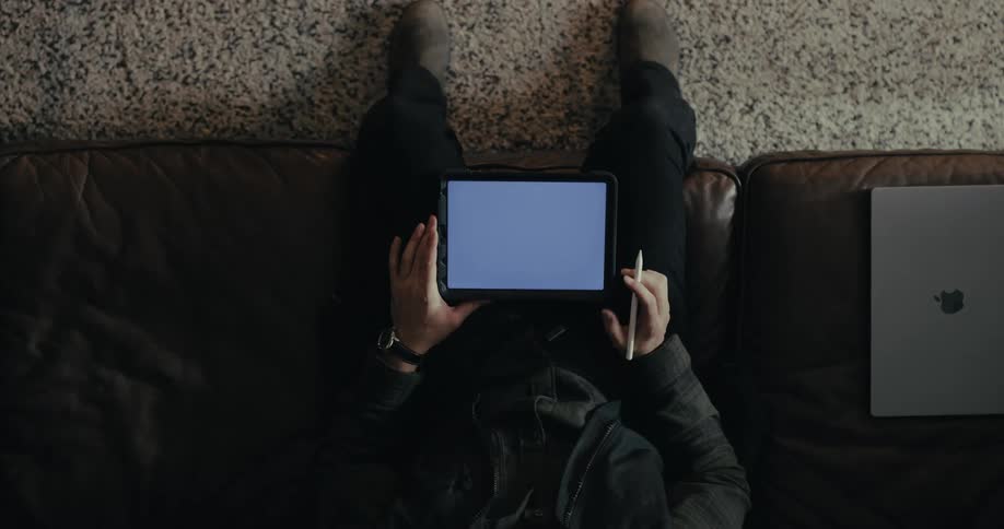 A person is sitting on a couch holding a tablet with a blank screen in one hand and a stylus in the other with a laptop placed beside them