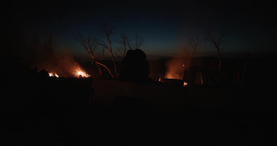 Man and woman sitting in the back of a truck kiss while silhouetted against multiple bonfires at night