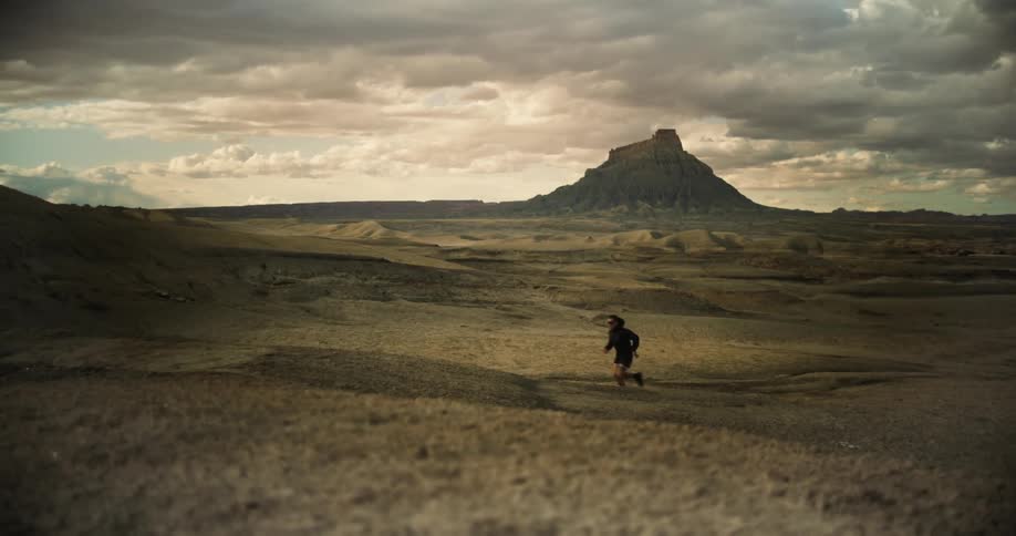 Person running on arid and desolate terrain with a rock formation in the background and a cloudy sky