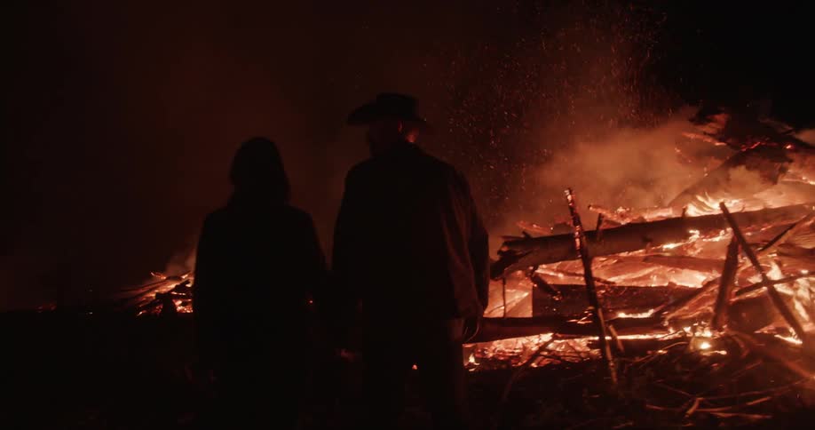 Two people holding hands are walking forward in a night environment with several bonfires burning wooden logs