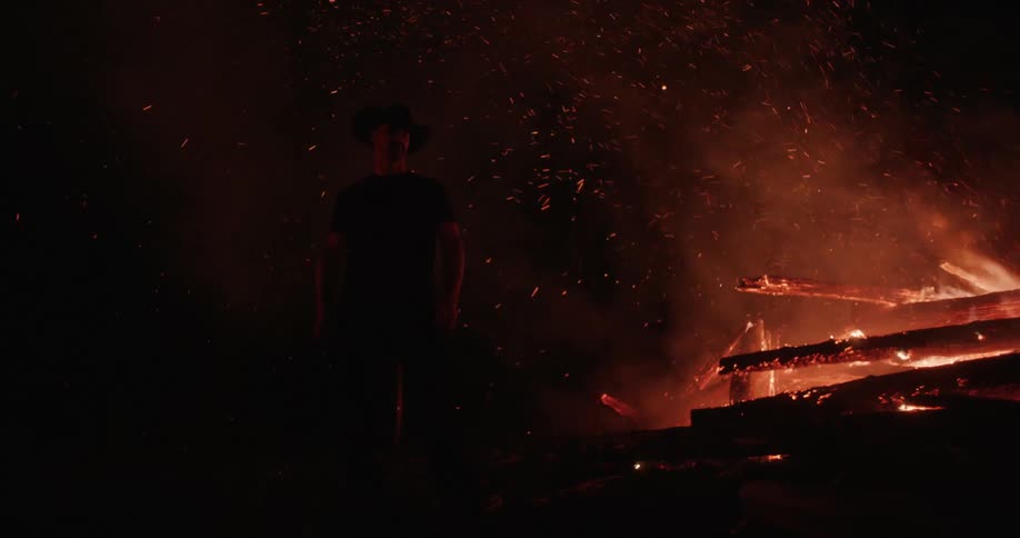 Man stands close to a smoking bonfire at night