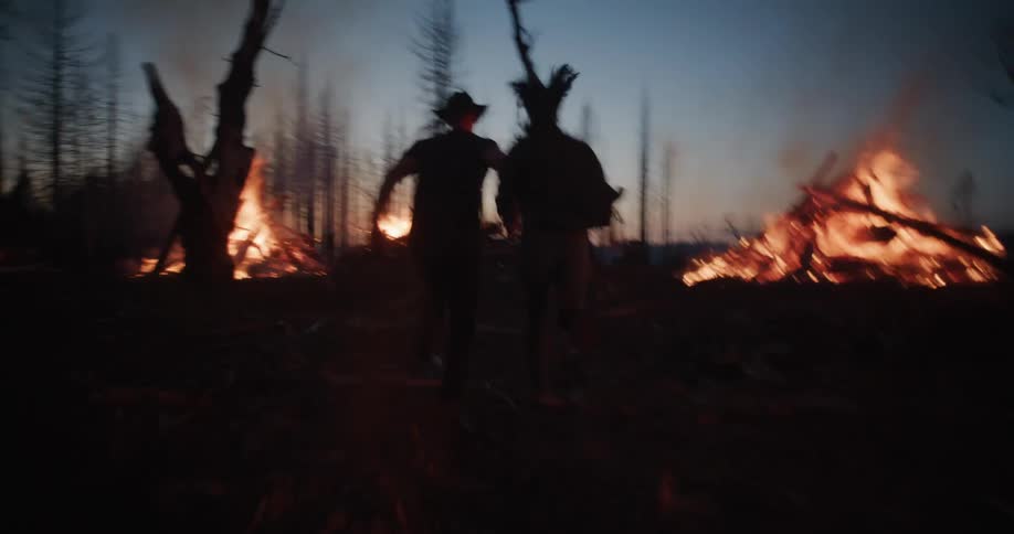 Two silhouetted figures a man wearing a cowboy hat and a woman hold hands as they run through a desolate landscape of burnt trees and smoldering fires