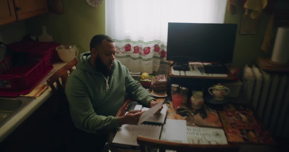 A person in a green hoodie sits at a kitchen table with a notebook pen and various papers appearing to work on taxes