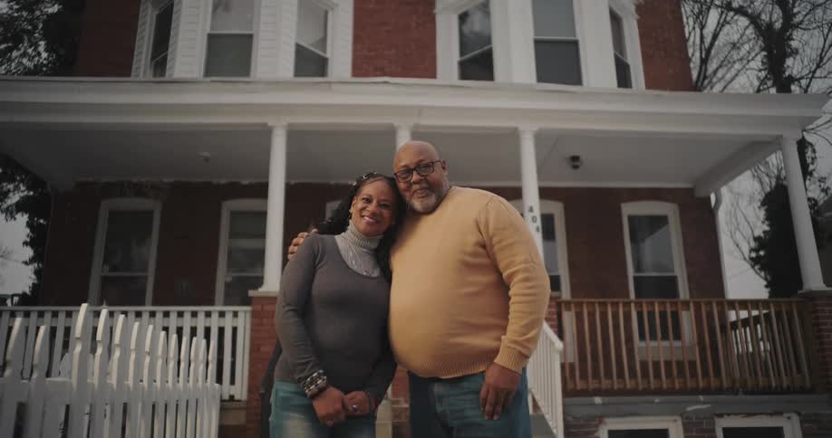 A man and a woman are standing in front of a twostory brick house with a white picket fence and a wooden porch