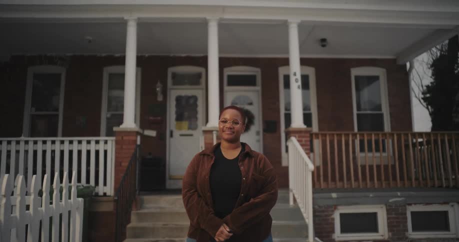 A woman standing in front of a twostory brick house with a white picket fence and a wooden porch