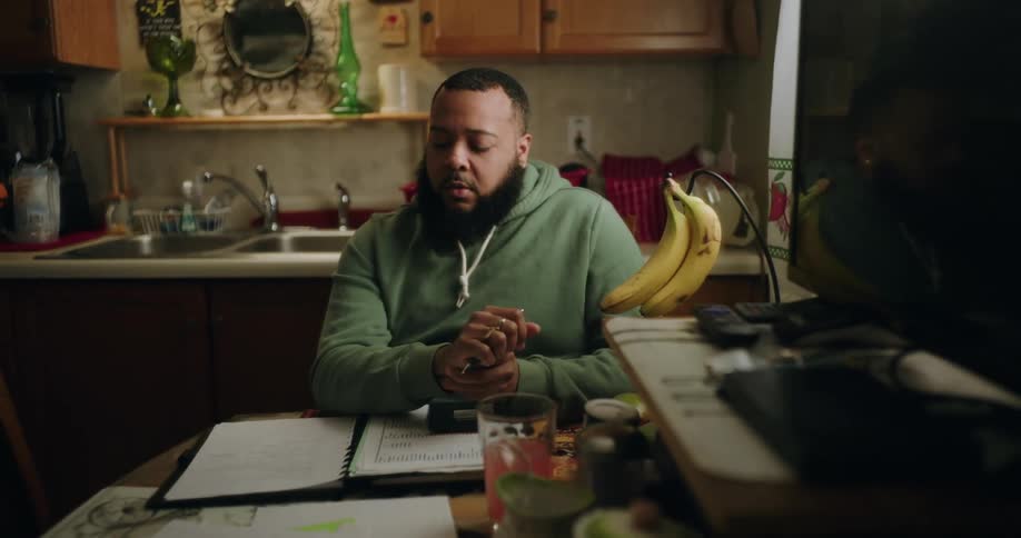 A person in a green hoodie sits at a kitchen table with a notebook pen and various papers appearing to work on taxes