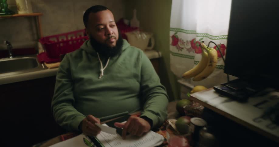 A person in a green hoodie sits at a kitchen table with a notebook pen and various papers appearing to work on taxes