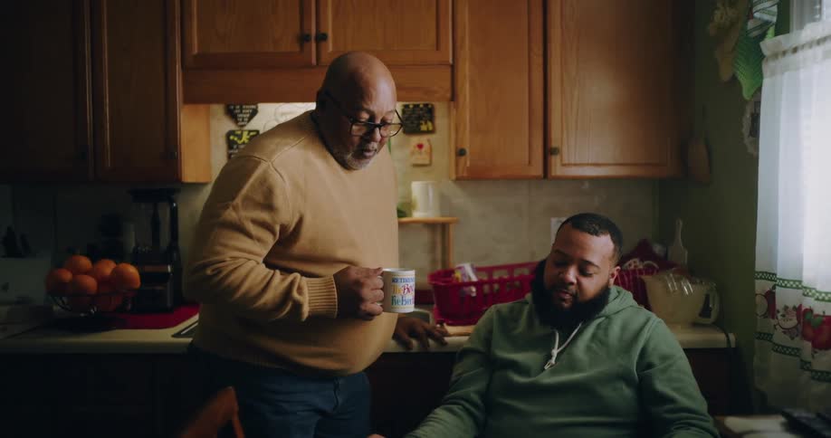 Two men in the kitchen with one of them sitting talking