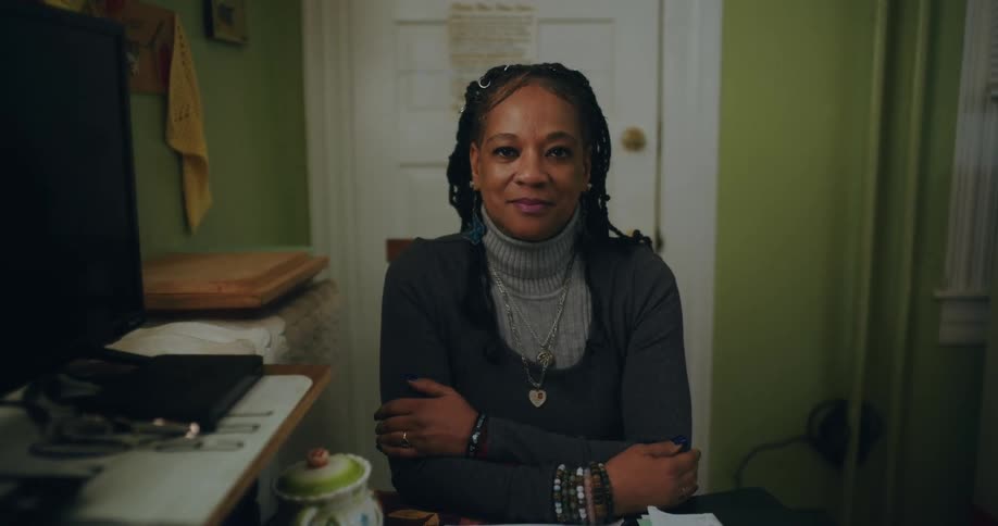 The face of a woman staring straight ahead with her arms crossed in a room with a white door in the background