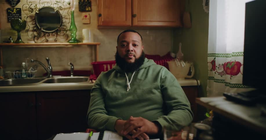 A bearded african american man in a green hoodie sits at a table in a kitchen a monitor sink and a window are visible