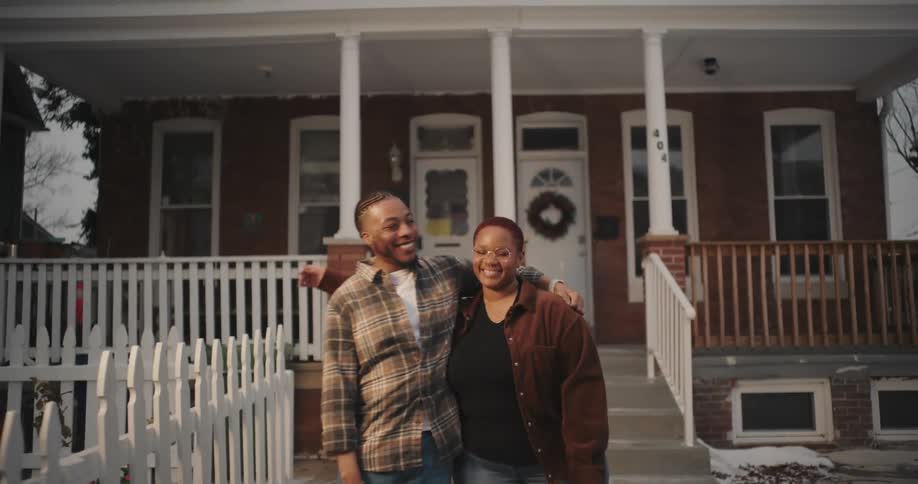 A man and a woman are standing in front of a twostory brick house with a white picket fence and a wooden porch