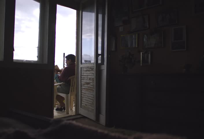 A silhouetted figure sits reading on a bright porch as seen from a dimly lit interior room adorned with framed pictures