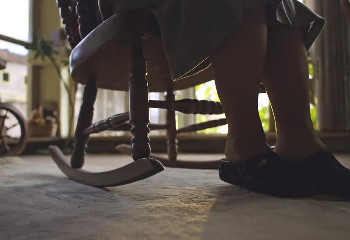 A closeup view of a persons legs and feet wearing dark slippers sitting on a wooden rocking chair in a sunlit room with a carpeted floor and a spinning wheel in the background