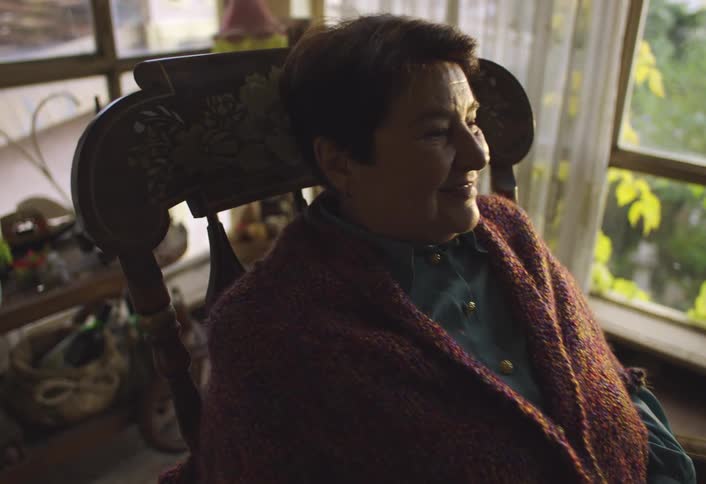 A woman wrapped in a thick brown knitted blanket sits in a rocking chair in a cozy room near a window