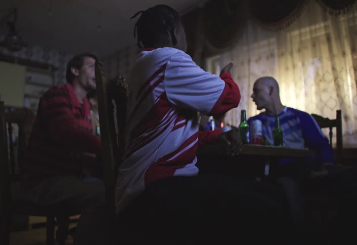 Friends are sitting around a table in a dimly lit room with bottles of beer on the table watching sports on tv