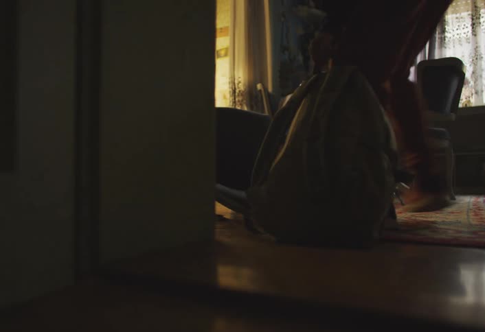 A teenage boy knocks over a chair while grabbing a backpack in a dim dining room picks it up with a womans help and then runs away