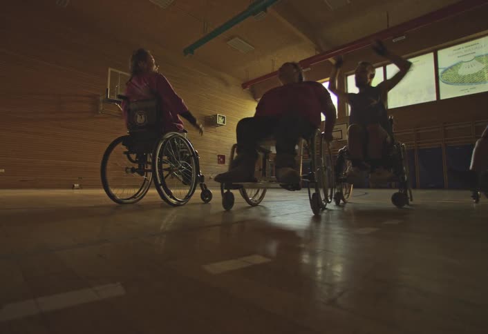 People in wheelchairs play basketball in an indoor gymnasium with sunlight streaming through large windows