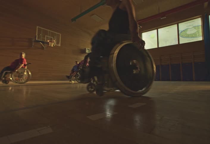 Teams in wheelchairs play basketball in an indoor gymnasium with sunlight streaming through large windows