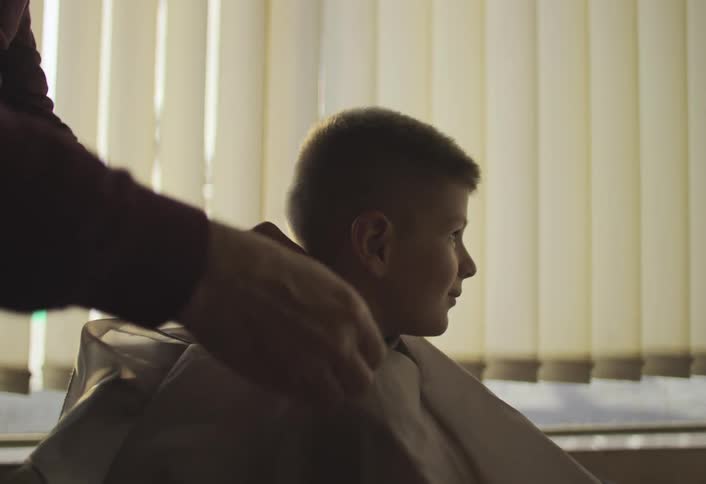 Seated boy smiles as a barber puts on an apron