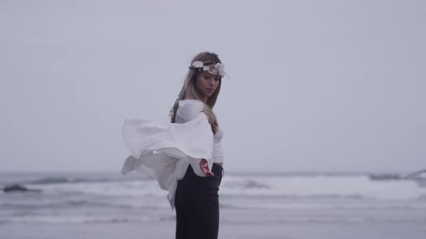 Woman wearing a flower crown and a blouse with fluttering sleeves is standing on the beach with the sea in the background