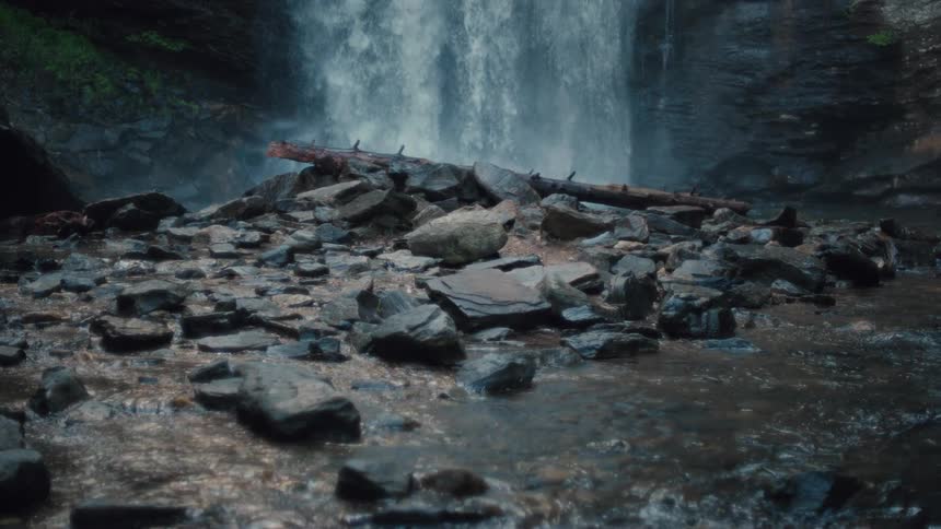 A cascading waterfall pours over rocky ledges into a shallow stream with a fallen log resting among scattered stones in the foreground
